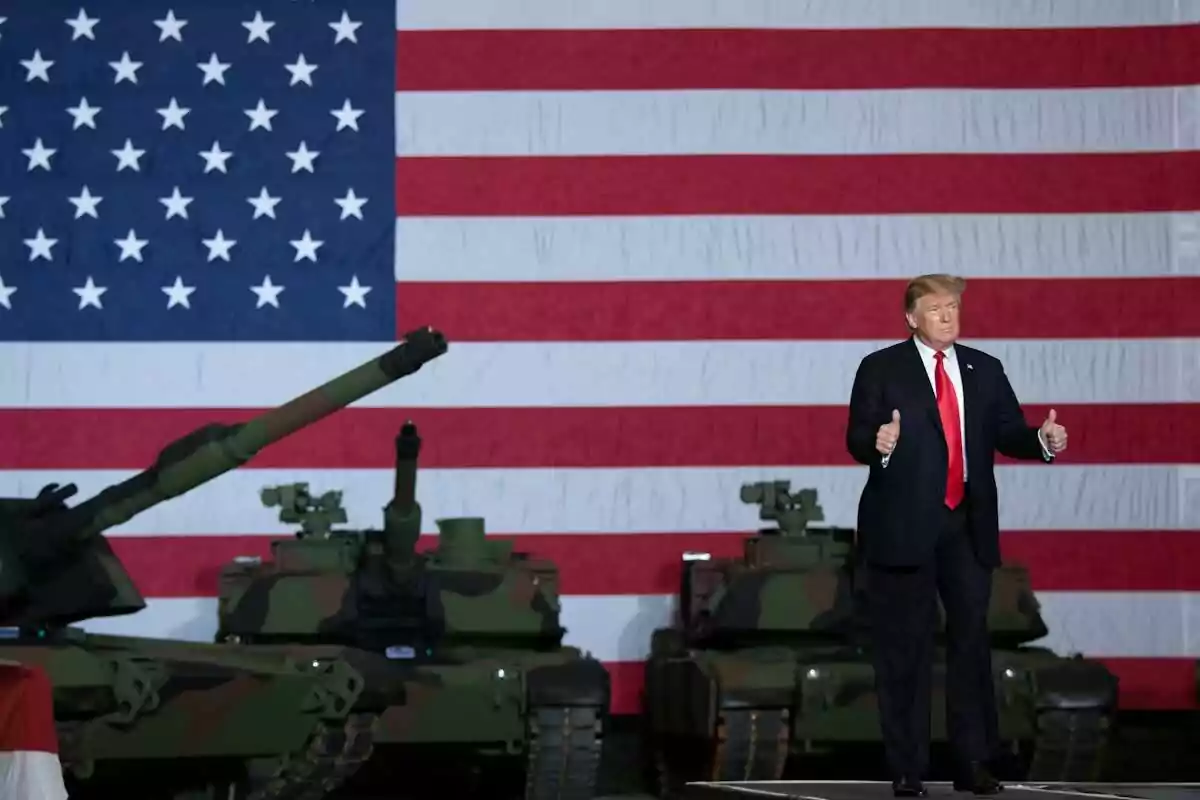 Man in a suit with a red tie giving a thumbs-up gesture in front of military tanks and a large United States flag Man in a suit with a red tie giving a thumbs-up gesture in front of military tanks and a large United States flag