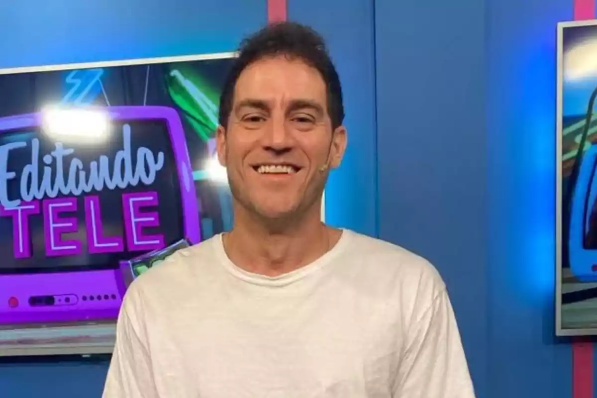 Un hombre sonriente con camiseta blanca frente a un fondo de estudio de televisión con pantallas que muestran gráficos coloridos.