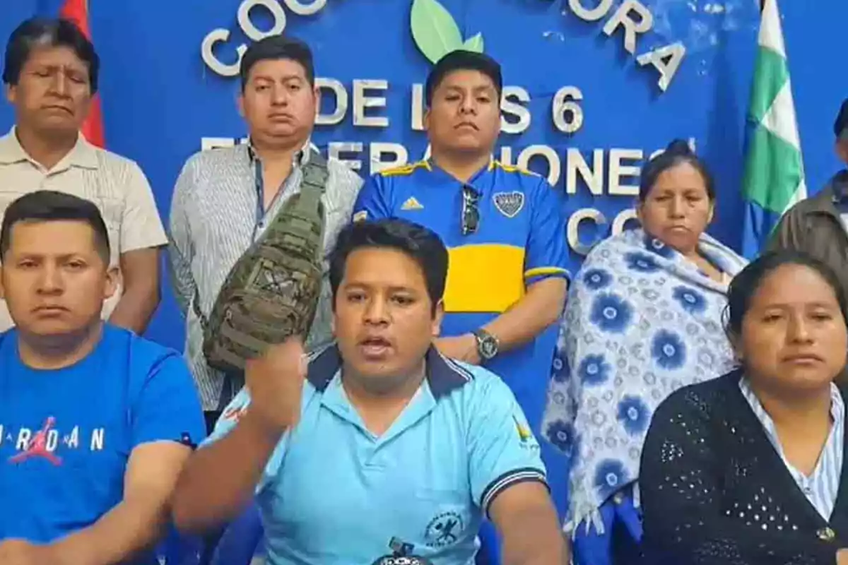 A group of people is sitting and standing in front of a blue background with white letters; some of them look serious, and a person in the center appears to be speaking or making a gesture with their hand.