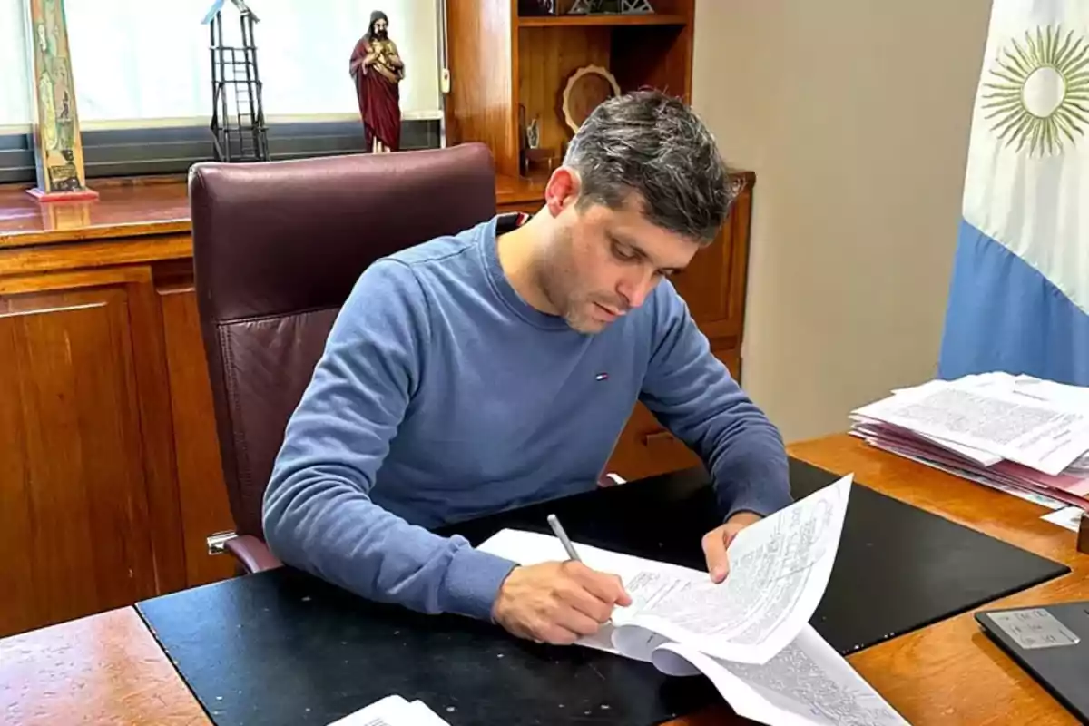 Un hombre sentado en un escritorio revisando y firmando documentos, con una bandera argentina y una figura religiosa en el fondo.