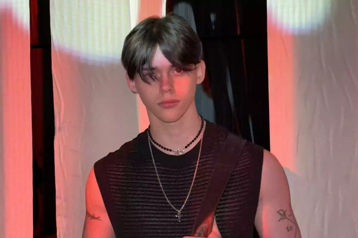 Un joven con cabello oscuro y liso, vistiendo una camiseta sin mangas y collares, está de pie frente a un fondo iluminado con luces rojas y blancas.