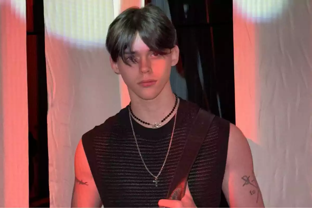 Un joven con cabello oscuro y liso lleva una camiseta sin mangas negra y collares, posando frente a un fondo iluminado en tonos cálidos.