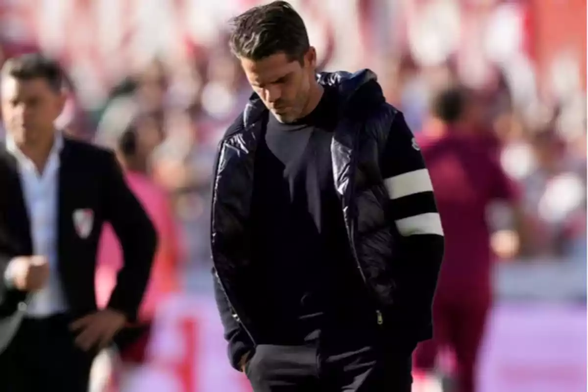 A man in a dark jacket with white stripes on the sleeves walks with his head down in a sports setting. A man in a dark jacket with white stripes on the sleeves walks with his head down in a sports setting.