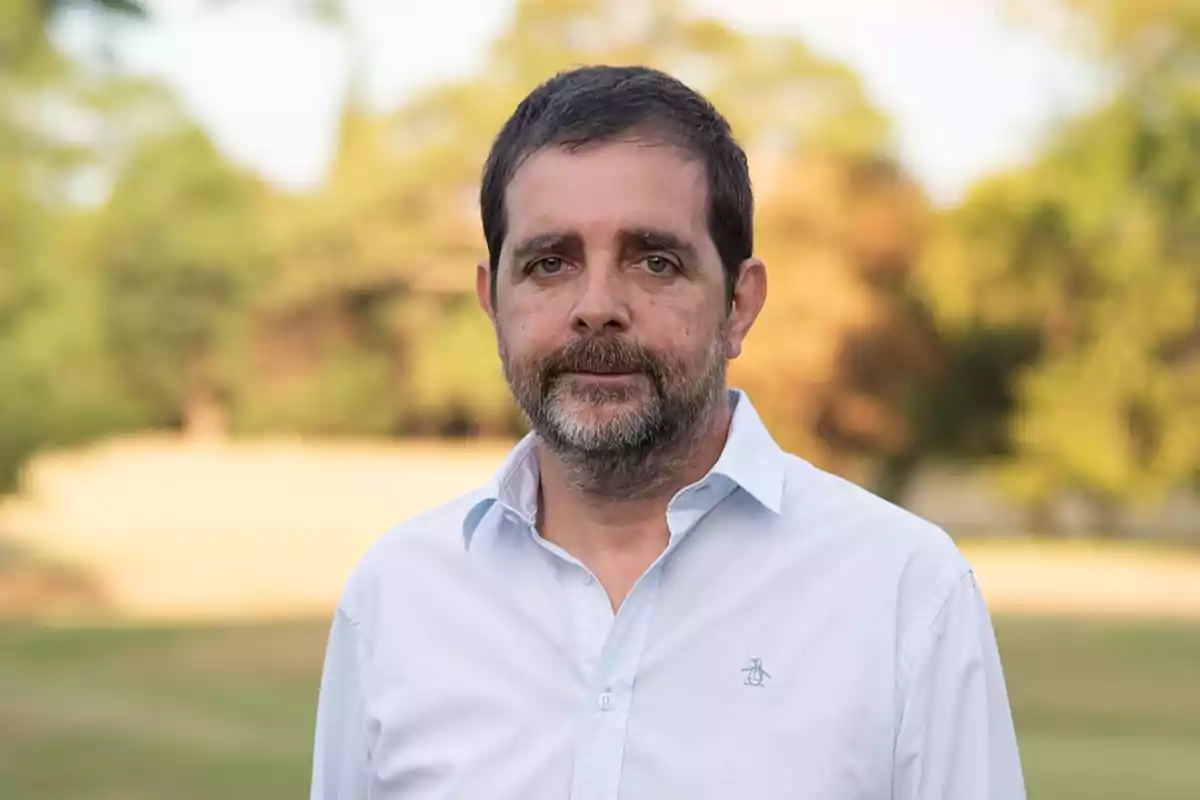 Hombre con barba y camisa blanca de pie en un entorno natural con árboles desenfocados al fondo.