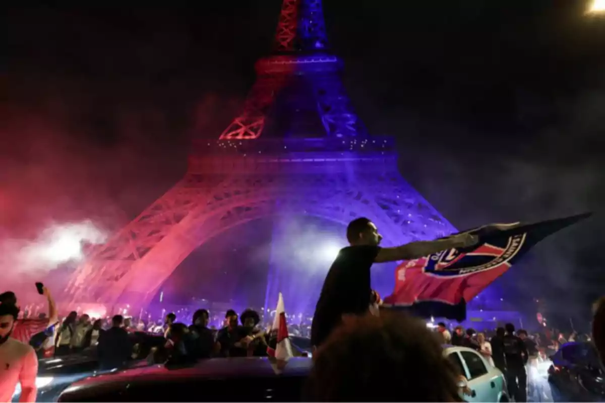Nighttime celebration with a crowd in front of the Eiffel Tower lit up in blue and red as someone waves a PSG flag. Nighttime celebration with a crowd in front of the Eiffel Tower lit up in blue and red as someone waves a PSG flag.