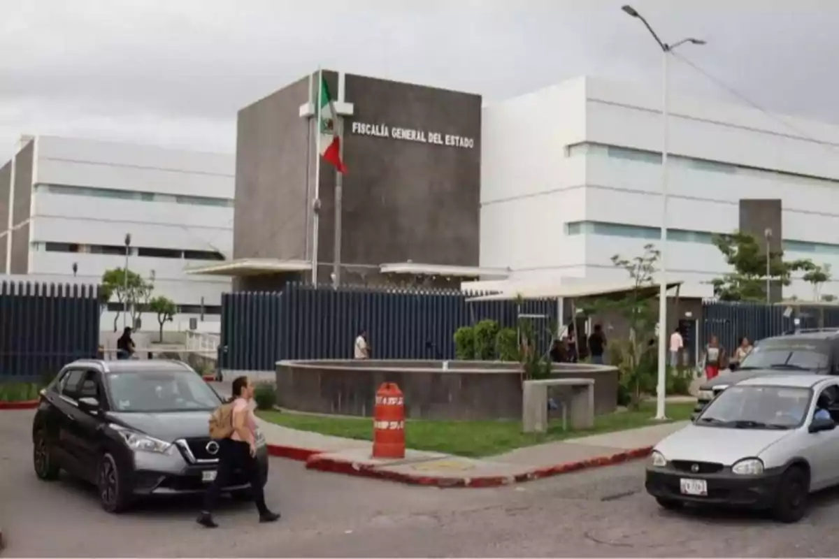 Edificio moderno de la Fiscalía General del Estado con bandera de México al frente y varios vehículos y personas en la calle.