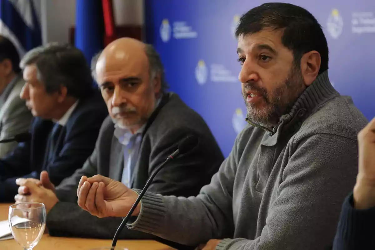 Un grupo de hombres sentados en una mesa durante una conferencia de prensa, uno de ellos hablando frente a un micrófono con fondo azul institucional.