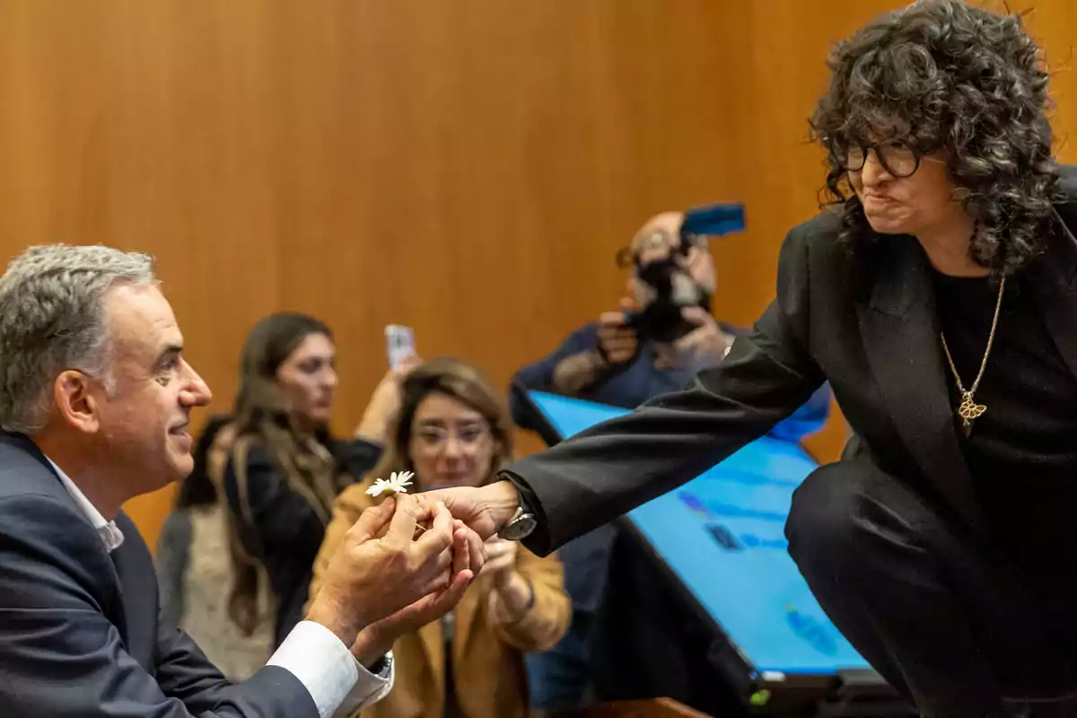 A man hands a flower to a woman at an event while several people watch and take photos. A man hands a flower to a woman at an event while several people watch and take photos.