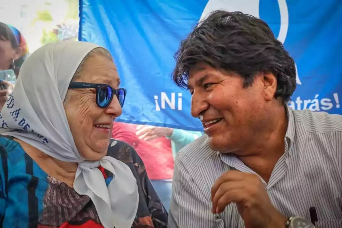 Dos personas sonríen mientras conversan frente a una bandera azul.