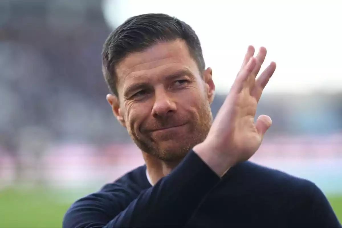 Short-haired man with a trimmed beard waving and slightly smiling in a blurred outdoor setting