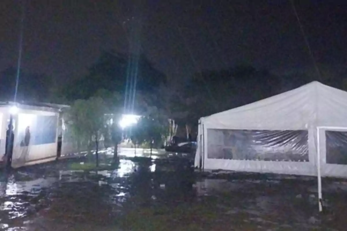 Carpas blancas instaladas en un terreno mojado durante la noche con charcos de agua reflejando las luces Carpas blancas instaladas en un terreno mojado durante la noche con charcos de agua reflejando las luces