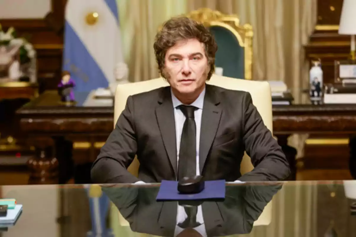 Un hombre con traje oscuro y corbata negra está sentado en un escritorio de oficina elegante con una bandera argentina al fondo.