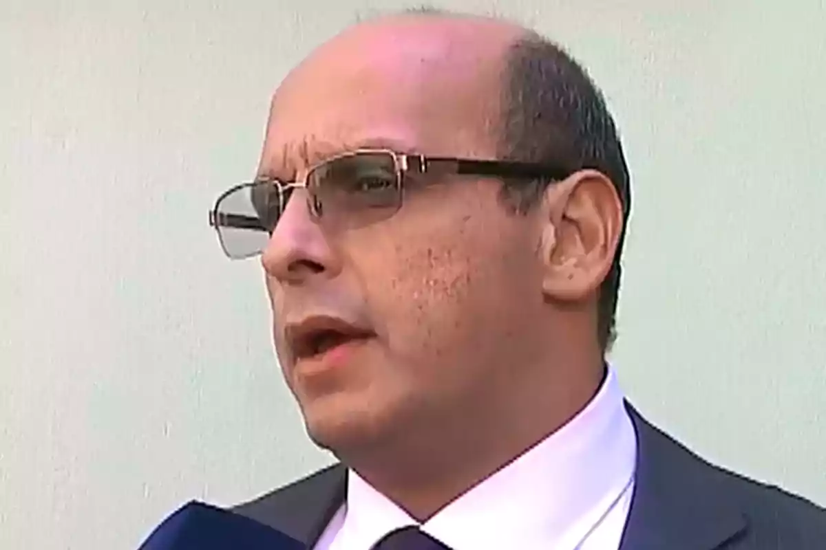 Hombre calvo con gafas y traje oscuro hablando frente a una pared clara Hombre calvo con gafas y traje oscuro hablando frente a una pared clara