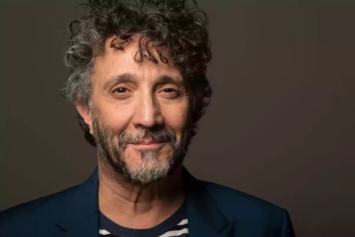 A man with curly hair and a beard slightly smiles while wearing a blue jacket and a striped jersey against a dark background.