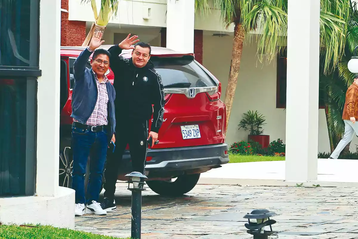 Two men smile and wave in front of a red truck parked in a driveway surrounded by palm trees and plants.
