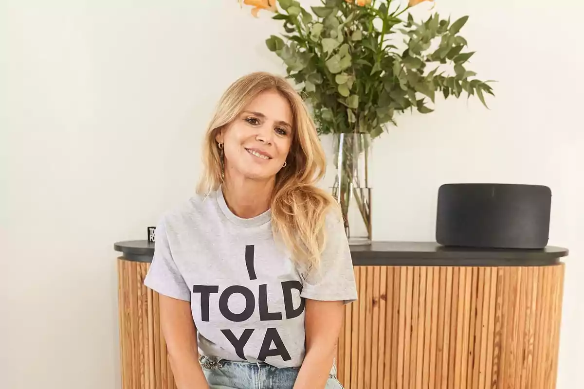 Mujer sonriendo con camiseta gris que dice "I TOLD YA" sentada frente a un mueble de madera con un jarrón de flores y un altavoz.