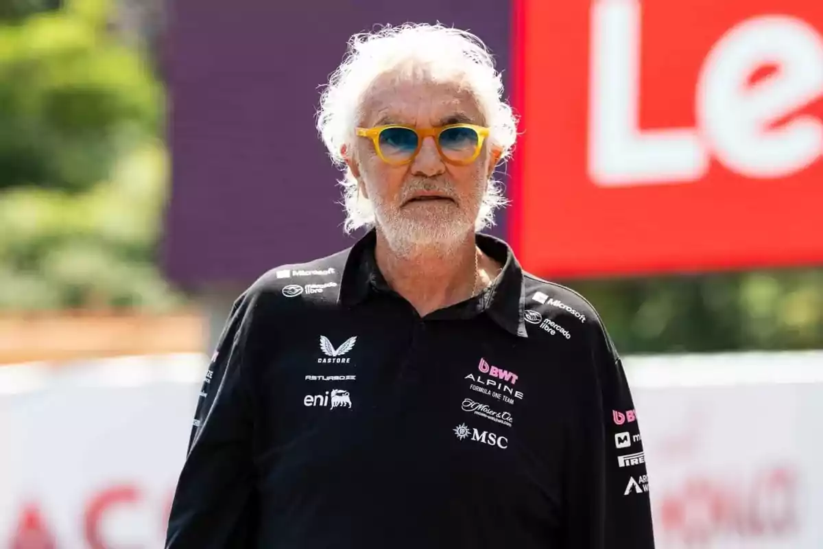 Gray-haired man wearing yellow-framed sunglasses and a black Alpine Formula 1 team jersey in an outdoor setting