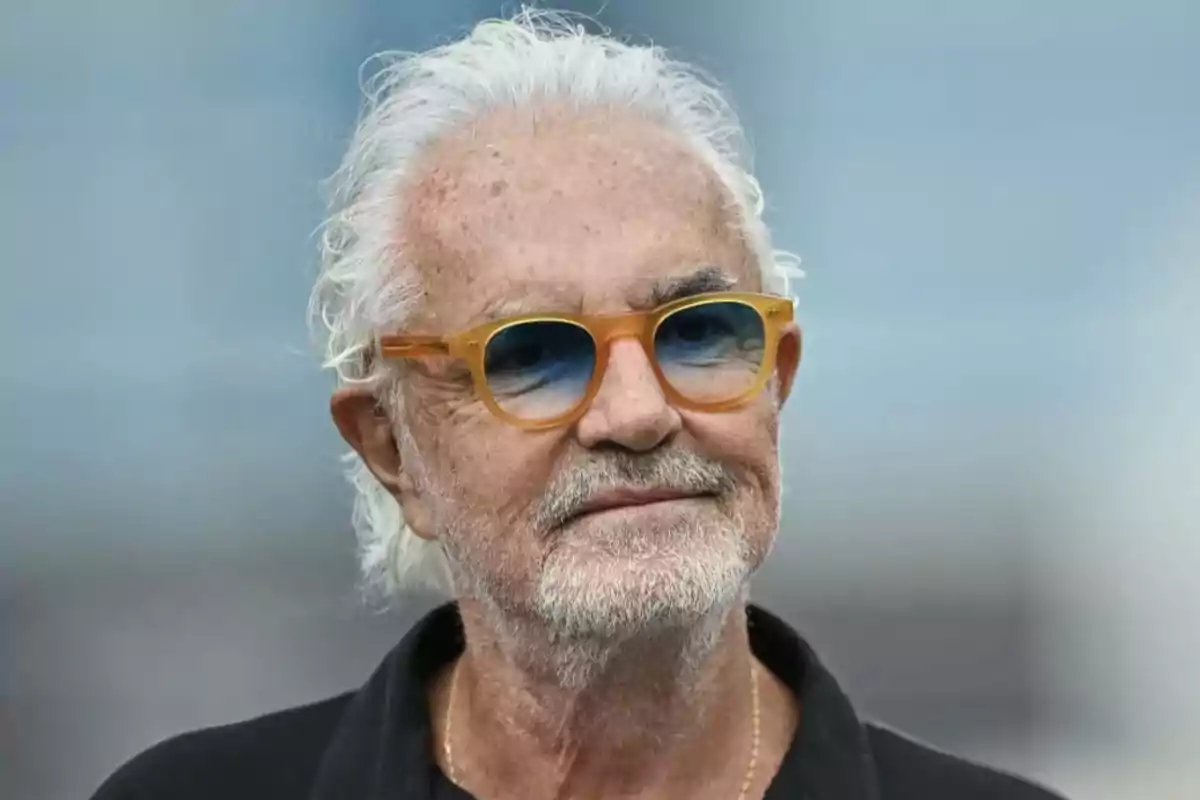 An older man with white hair and orange sunglasses, wearing a dark jersey, with a blurred background. An older man with white hair and orange sunglasses, wearing a dark jersey, with a blurred background.