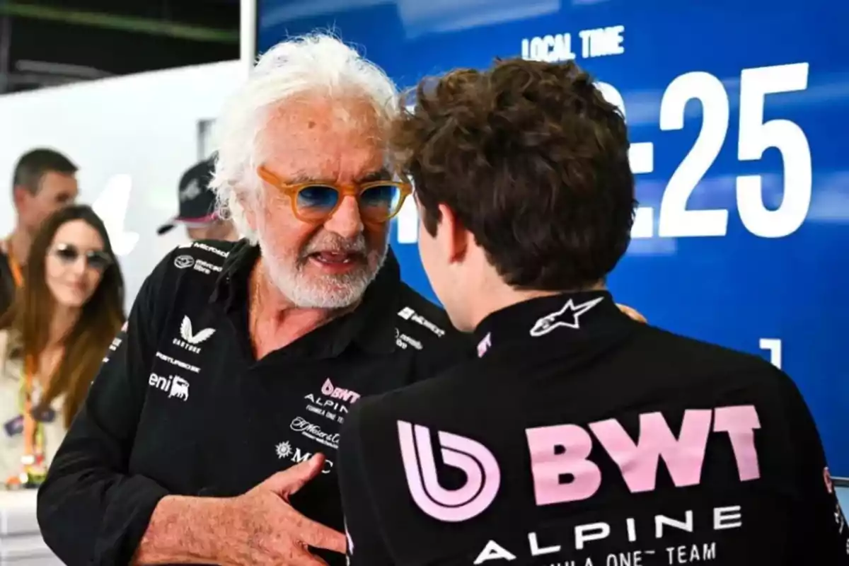 Un hombre de cabello blanco y gafas de sol habla con un joven vestido con uniforme del equipo Alpine de Fórmula 1 frente a un fondo azul con números grandes blancos