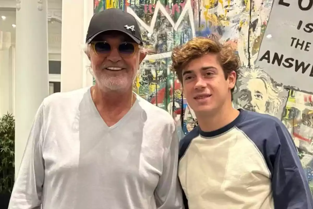 Two people pose together in front of a colorful artwork in an indoor setting.