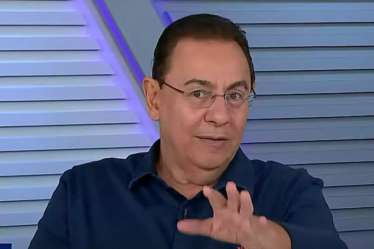Un hombre con gafas y camisa oscura gesticulando frente a un fondo con paneles. Un hombre con gafas y camisa oscura gesticulando frente a un fondo con paneles.