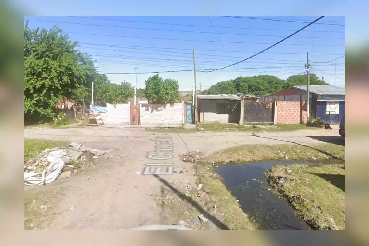 A dirt road with accumulated trash and brick and metal sheet houses in the background, surrounded by trees and electrical wires.