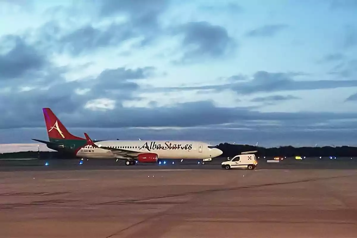 Un avión de la aerolínea AlbaStar en la pista de un aeropuerto al atardecer con un vehículo de servicio cercano.