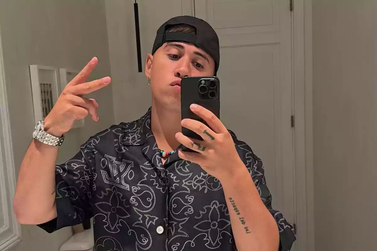 Un hombre joven con gorra negra y camisa estampada se toma una selfie en un espejo, haciendo un gesto de paz con la mano izquierda.