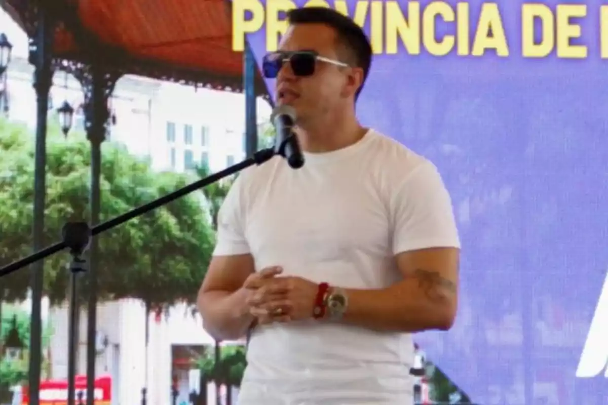 Un hombre con gafas de sol y camiseta blanca habla frente a un micrófono en un escenario al aire libre. Un hombre con gafas de sol y camiseta blanca habla frente a un micrófono en un escenario al aire libre.