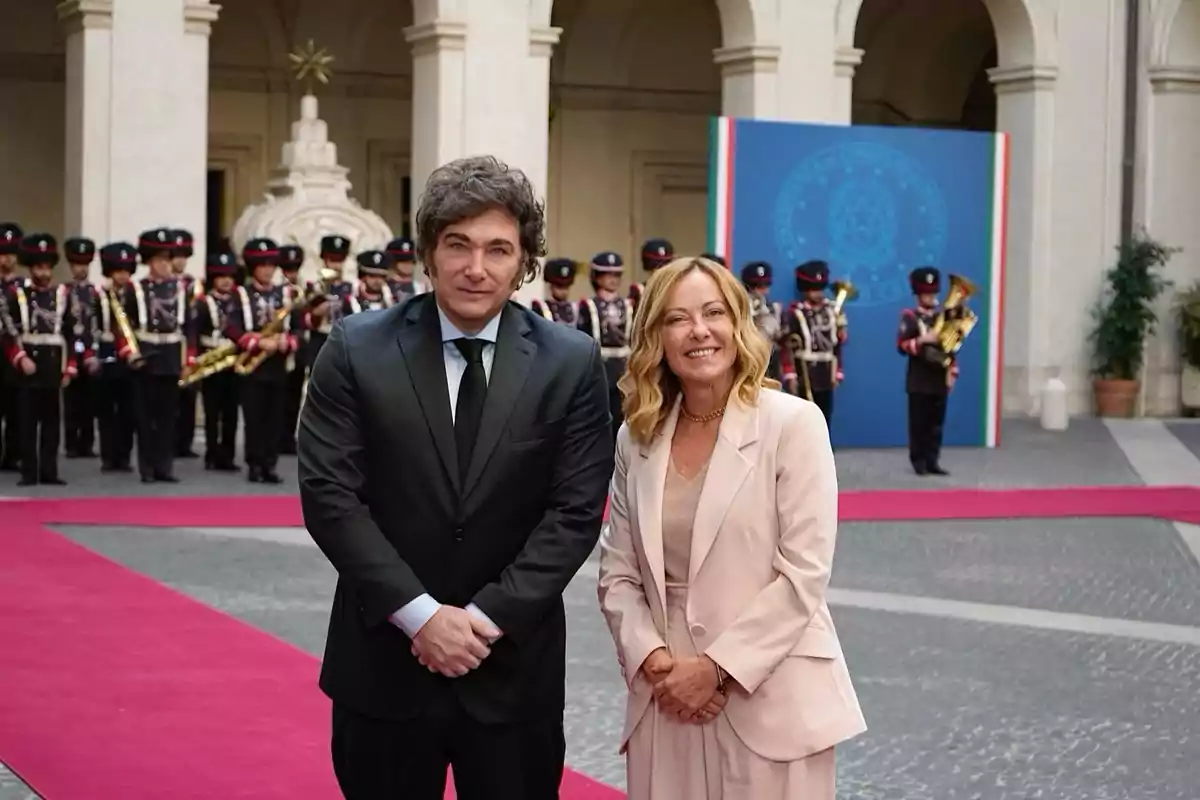 Dos personas posan sonrientes en un evento formal al aire libre con una banda militar uniformada al fondo y una alfombra roja sobre el suelo