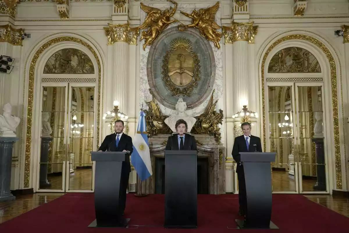 Tres personas de pie detrás de podios en una sala elegante con decoración dorada y una bandera de Argentina.