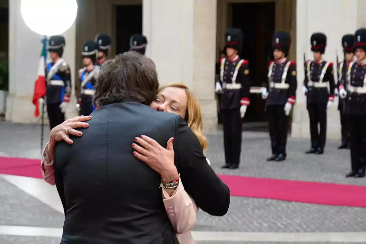 Dos personas se abrazan en primer plano mientras al fondo se observa una fila de guardias uniformados y una alfombra roja en un entorno formal Dos personas se abrazan en primer plano mientras al fondo se observa una fila de guardias uniformados y una alfombra roja en un entorno formal