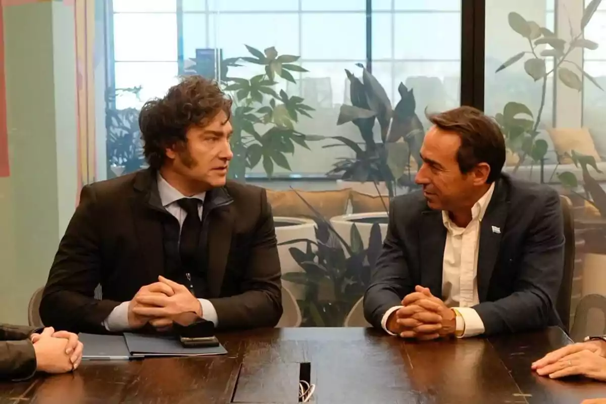 Dos hombres sentados en una mesa de madera, conversando en una oficina con plantas en el fondo.