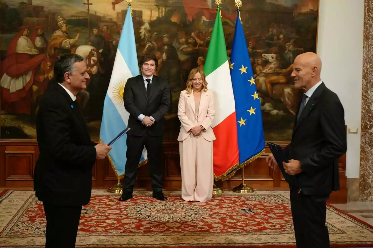 Cuatro personas elegantemente vestidas posan en una sala con alfombra roja y un gran cuadro de fondo, detrás de ellas se ven las banderas de Argentina, Italia y la Unión Europea.