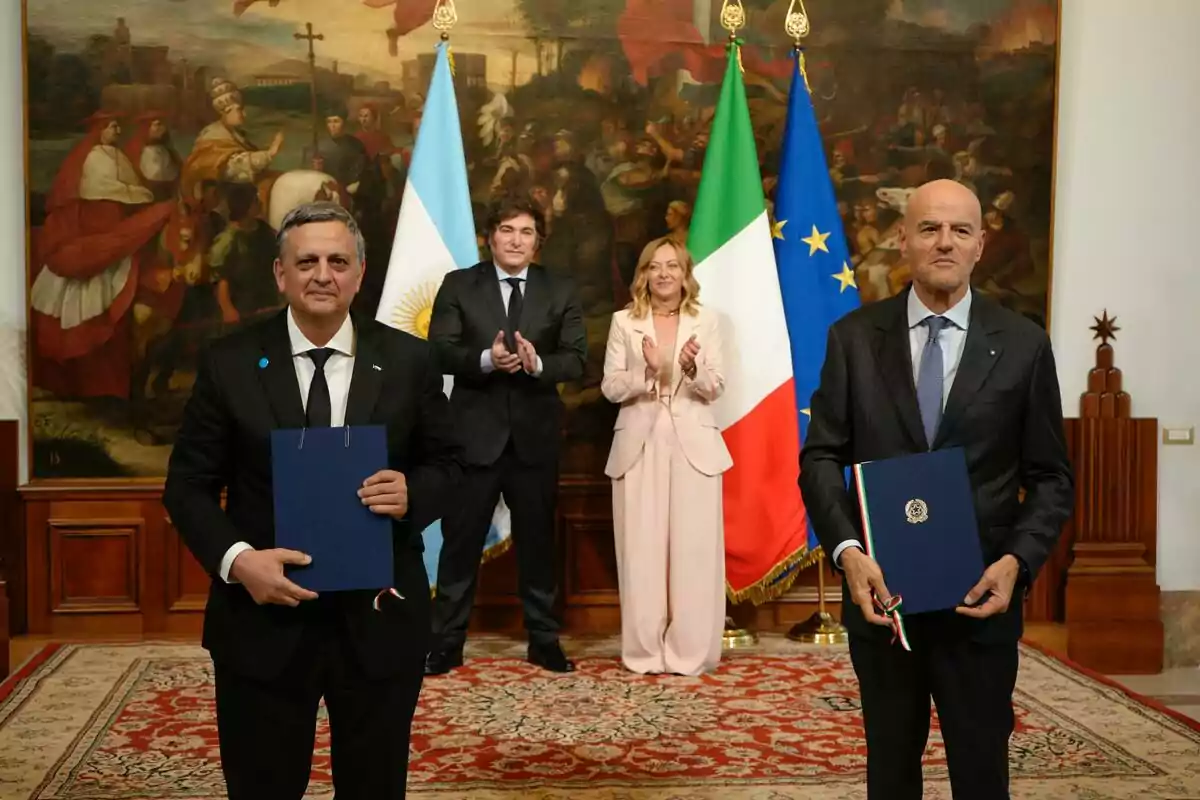Cinco personas posan en una sala elegante con alfombra y un gran cuadro al fondo, dos de ellas al frente sostienen carpetas azules y detrás se ven las banderas de Argentina, Italia y la Unión Europea.