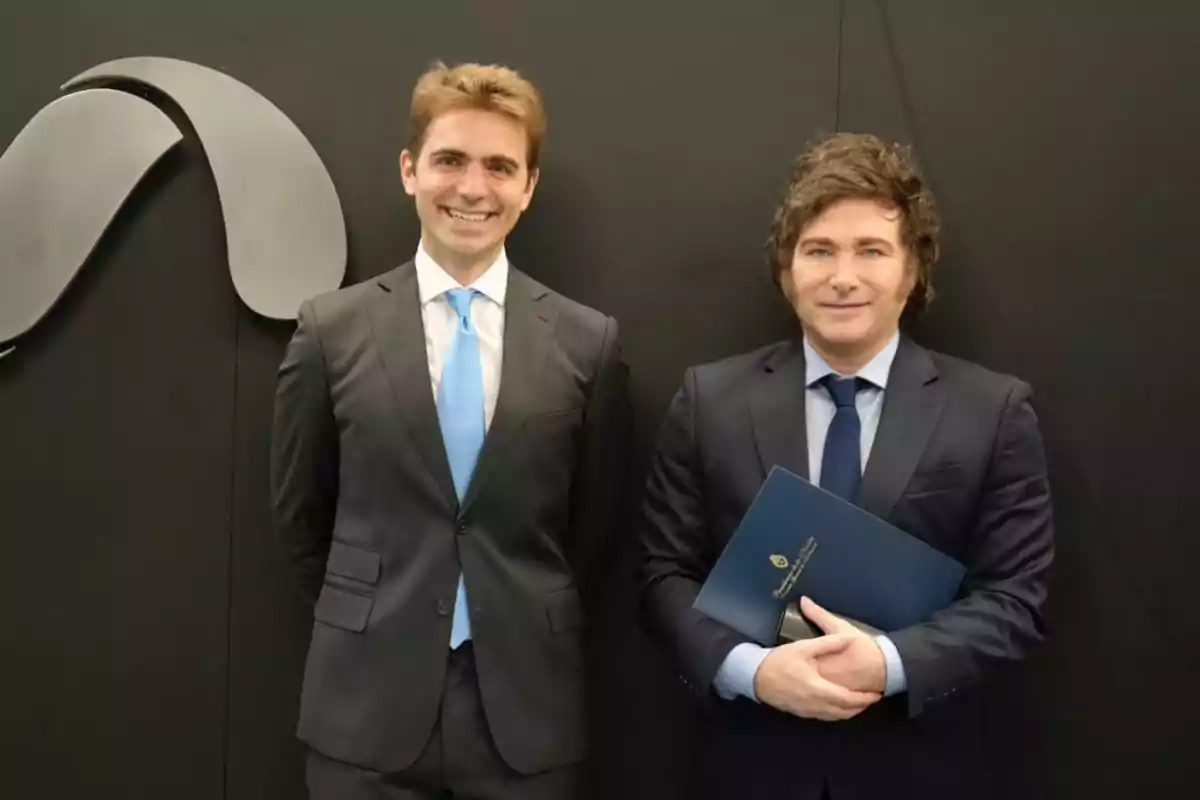 Dos hombres de traje posan frente a una pared negra con un logotipo abstracto.