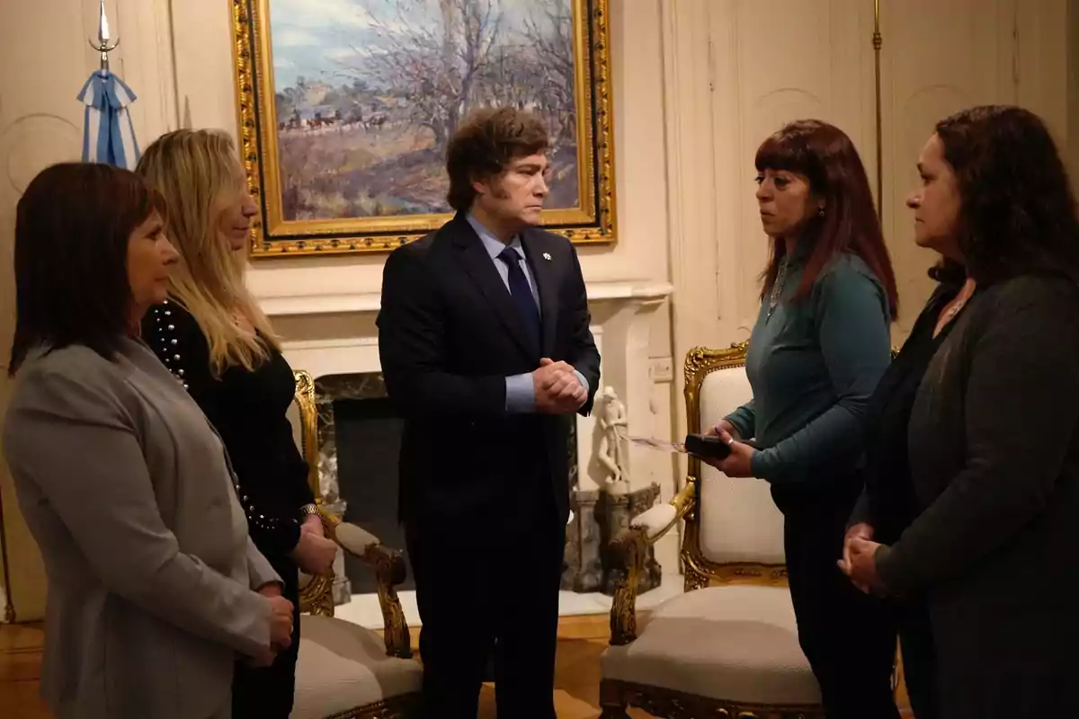 Un grupo de personas está reunido en una sala elegante con un cuadro y una bandera en el fondo, mientras conversan de pie alrededor de un hombre que lleva traje. Un grupo de personas está reunido en una sala elegante con un cuadro y una bandera en el fondo, mientras conversan de pie alrededor de un hombre que lleva traje.
