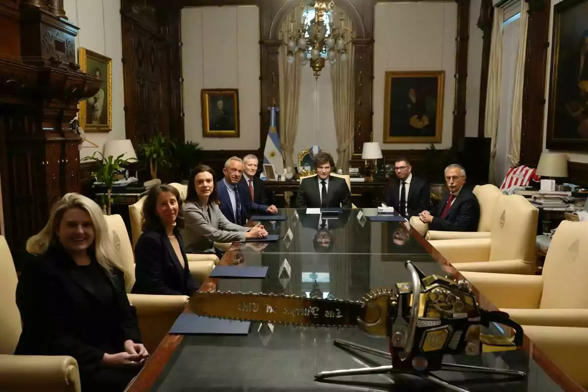 Ocho personas sentadas alrededor de una mesa de reuniones en una oficina elegante con una motosierra sobre la mesa y la bandera de Argentina al fondo Ocho personas sentadas alrededor de una mesa de reuniones en una oficina elegante con una motosierra sobre la mesa y la bandera de Argentina al fondo