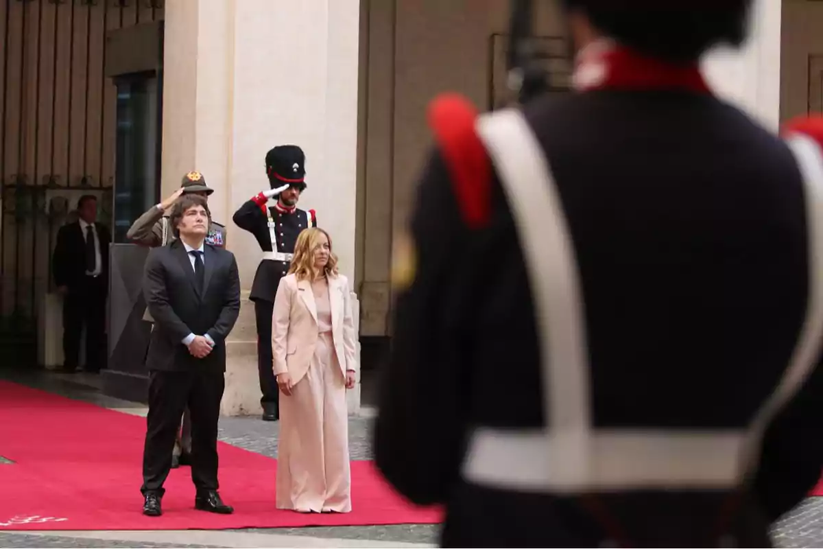 Dos personas de pie sobre una alfombra roja durante una ceremonia oficial con guardias uniformados al fondo y en primer plano Dos personas de pie sobre una alfombra roja durante una ceremonia oficial con guardias uniformados al fondo y en primer plano