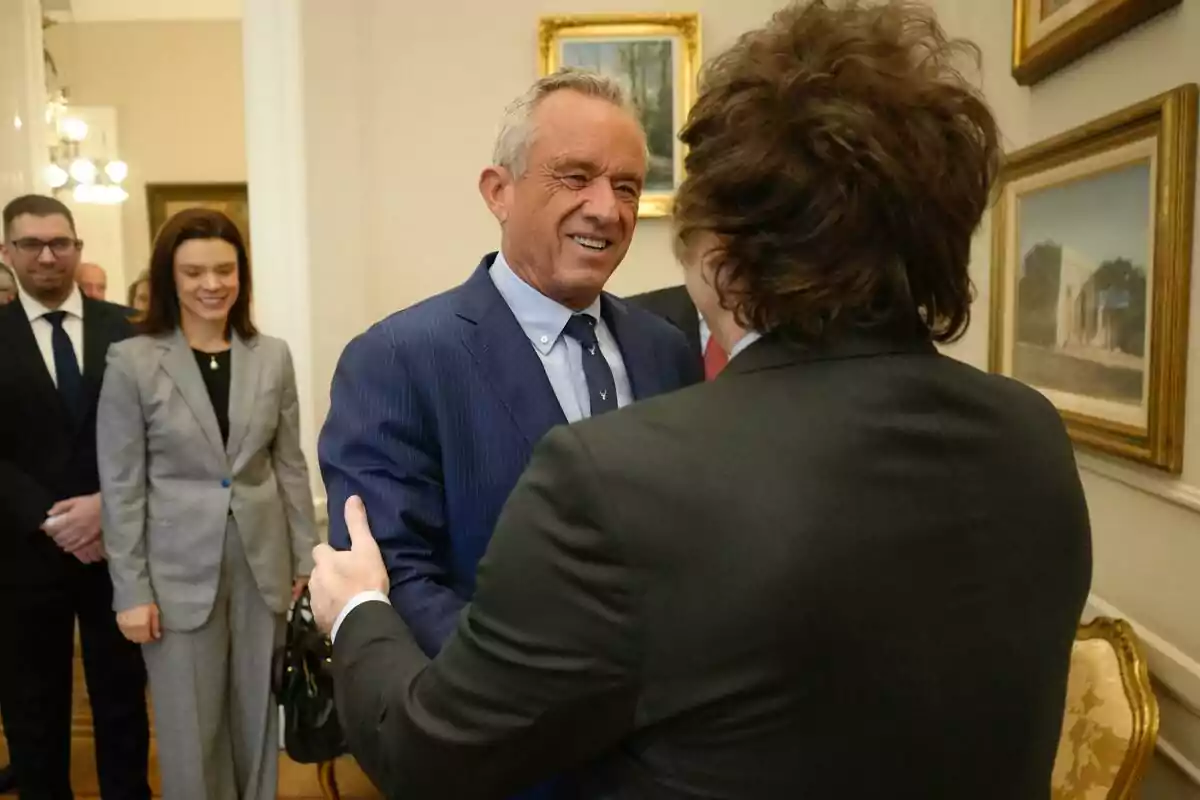 Dos hombres de traje se saludan sonrientes en una sala elegante mientras otras personas observan en el fondo Dos hombres de traje se saludan sonrientes en una sala elegante mientras otras personas observan en el fondo