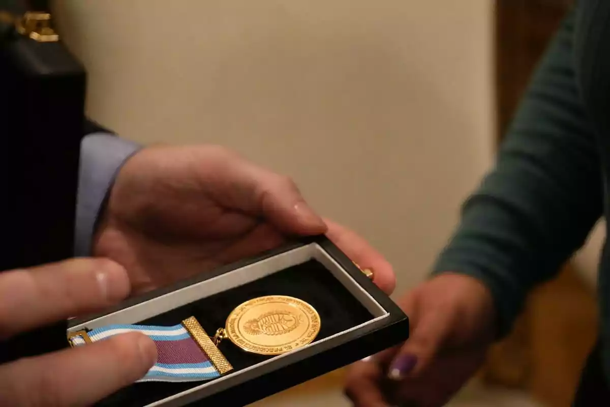 Una persona sostiene una caja que contiene una medalla dorada con una cinta azul y blanca. Una persona sostiene una caja que contiene una medalla dorada con una cinta azul y blanca.