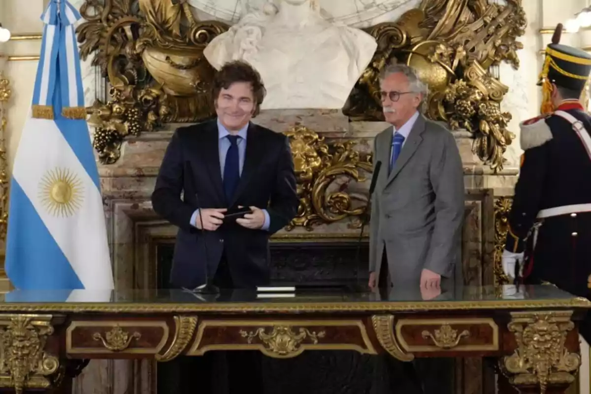 Dos hombres de pie junto a una mesa decorada con detalles dorados y una bandera argentina a la izquierda.