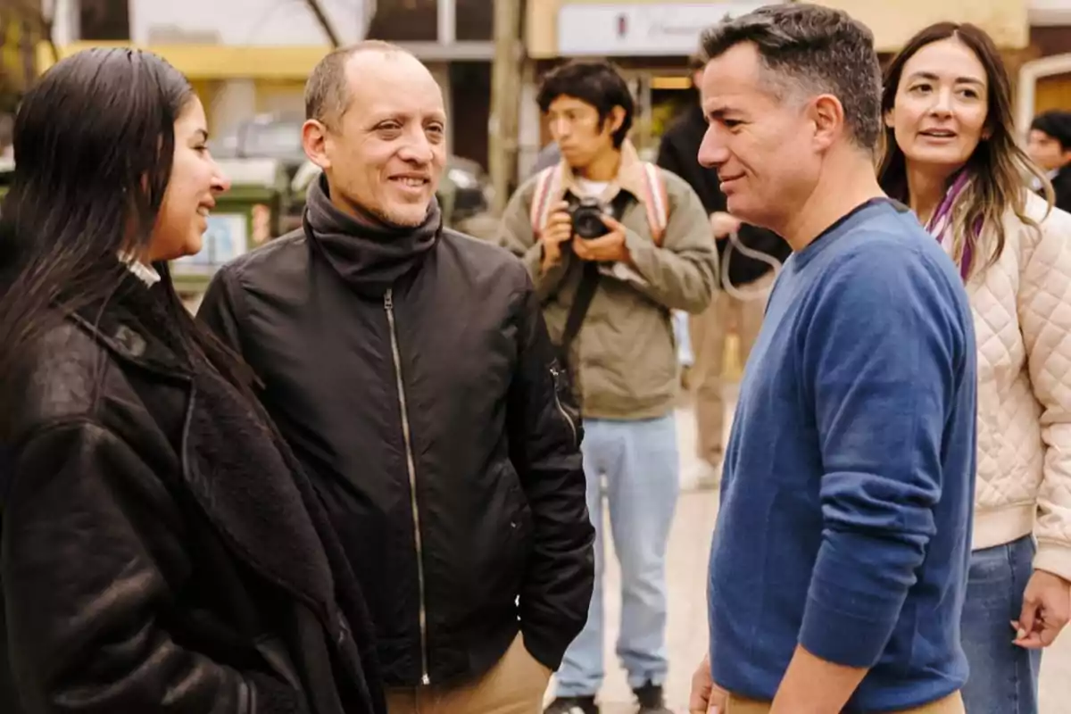 Varias personas conversan y sonríen al aire libre mientras un joven con cámara observa al fondo Varias personas conversan y sonríen al aire libre mientras un joven con cámara observa al fondo