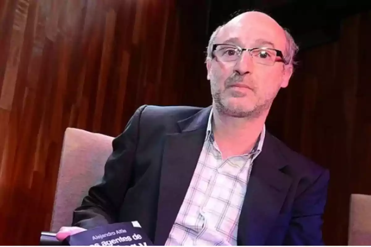 Un hombre con gafas y barba incipiente, vestido con una chaqueta oscura y camisa a cuadros, sostiene un libro mientras está sentado en una silla con un fondo de madera.