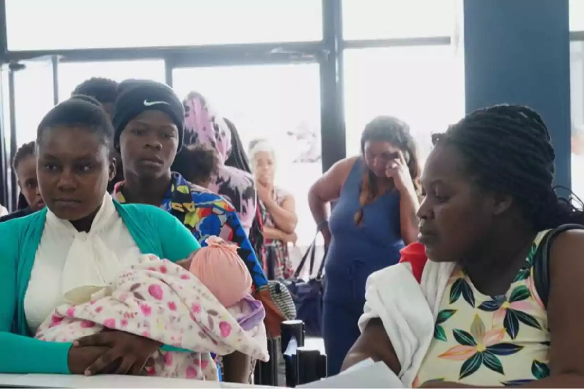 Varias personas, incluyendo una mujer con un bebé en brazos, esperan en una fila dentro de un edificio iluminado por la luz exterior que entra por las ventanas.