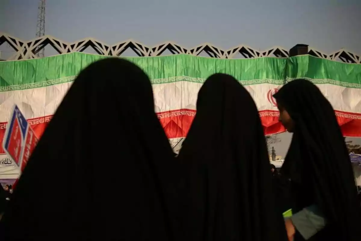 Mujeres vestidas con chador negro de espaldas frente a una gran bandera de Irán Mujeres vestidas con chador negro de espaldas frente a una gran bandera de Irán