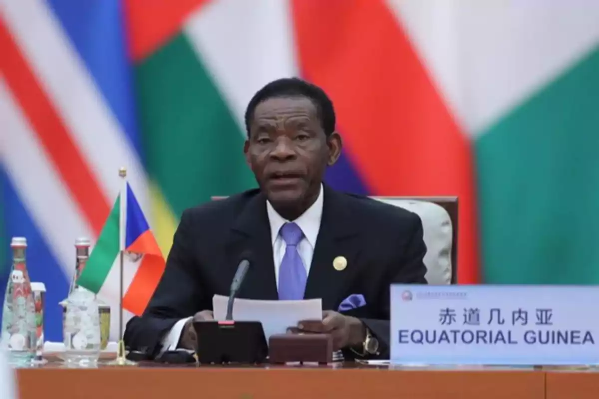 A man in a dark suit and blue tie is sitting at a conference table with a microphone, papers in his hand, and a nameplate that says Equatorial Guinea; in the background, flags of several countries can be seen. A man in a dark suit and blue tie is sitting at a conference table with a microphone, papers in his hand, and a nameplate that says Equatorial Guinea; in the background, flags of several countries can be seen.
