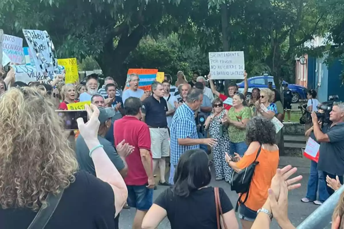 Un grupo de personas se manifiesta al aire libre sosteniendo carteles y aplaudiendo mientras una mujer graba la escena con su celular. Un grupo de personas se manifiesta al aire libre sosteniendo carteles y aplaudiendo mientras una mujer graba la escena con su celular.
