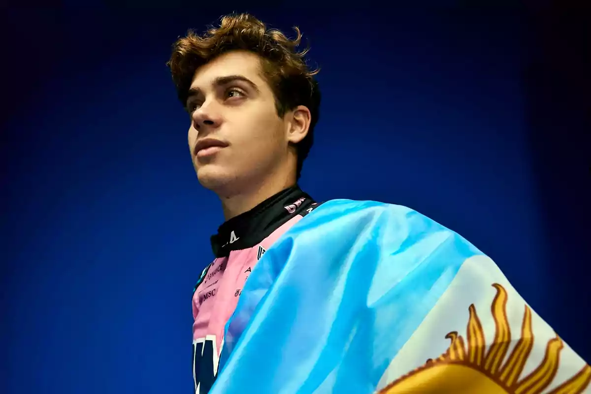 Un joven con una bandera argentina sobre los hombros, mirando hacia arriba con un fondo azul.