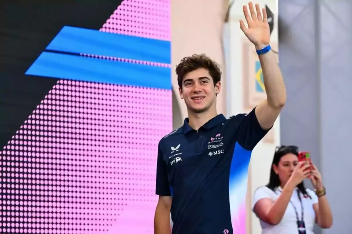 Young man in a blue t-jersey waves while a woman in the background takes a photo with her phone.